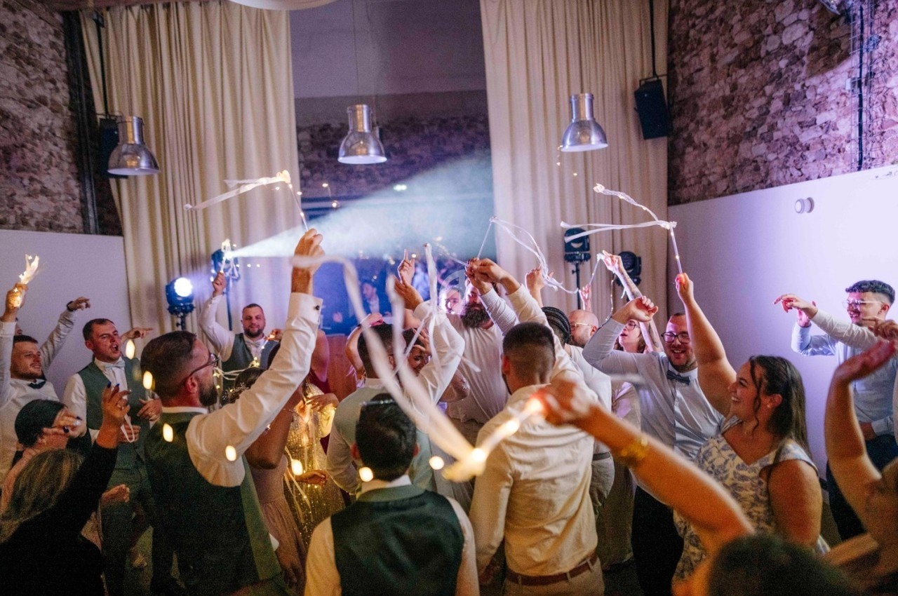 Des invités dansent et agitent des bâtons lumineux dans une salle décorée avec des murs en pierre et des rideaux blancs.