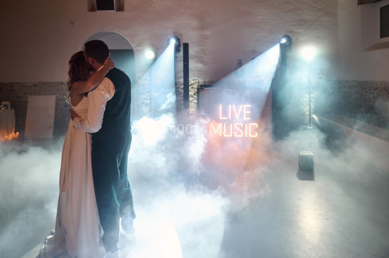 Un couple danse dans une salle éclairée par des projecteurs et de la fumée, avec un panneau ’Live Music’ en arrière-plan.