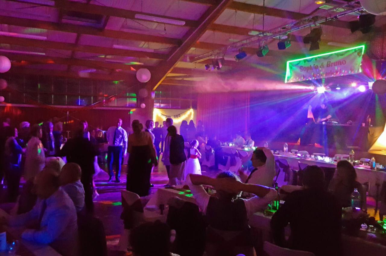 Des personnes dansent et discutent dans une salle de réception éclairée par des lumières colorées et un DJ sur scène.