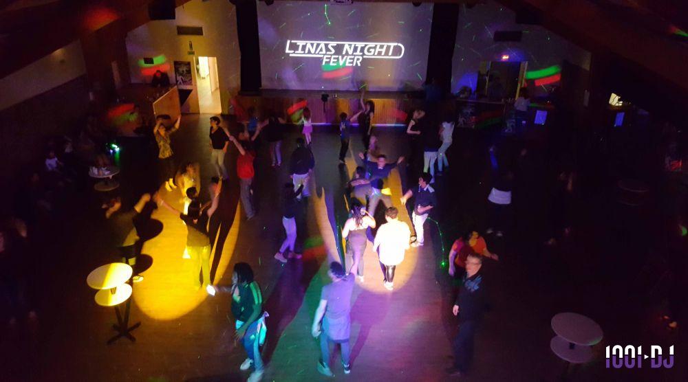 Personnes dansant dans une salle sombre éclairée par des lumières colorées, avec un écran affichant ’Linas Night Fever’.