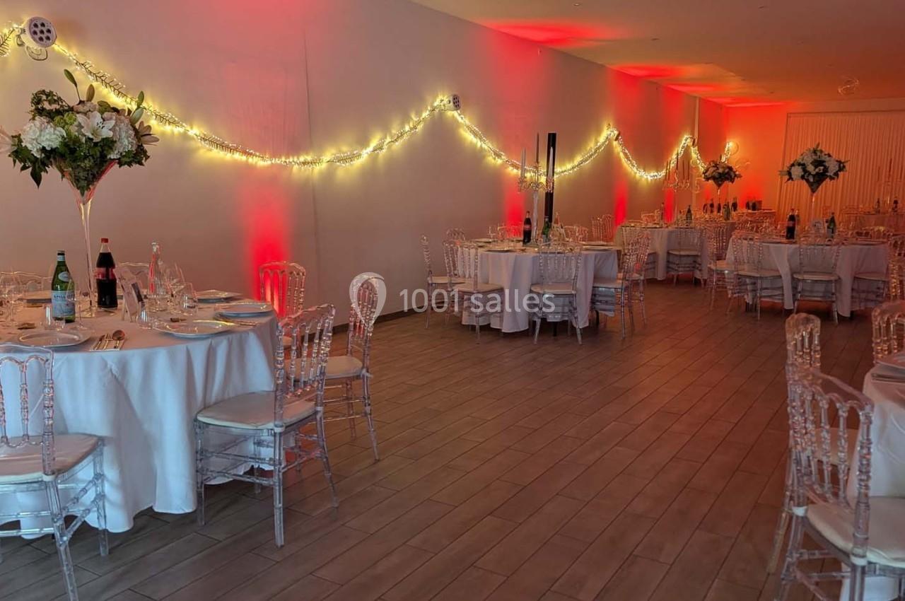 Salle de réception décorée avec des tables rondes, nappes blanches, chaises transparentes et guirlandes lumineuses.