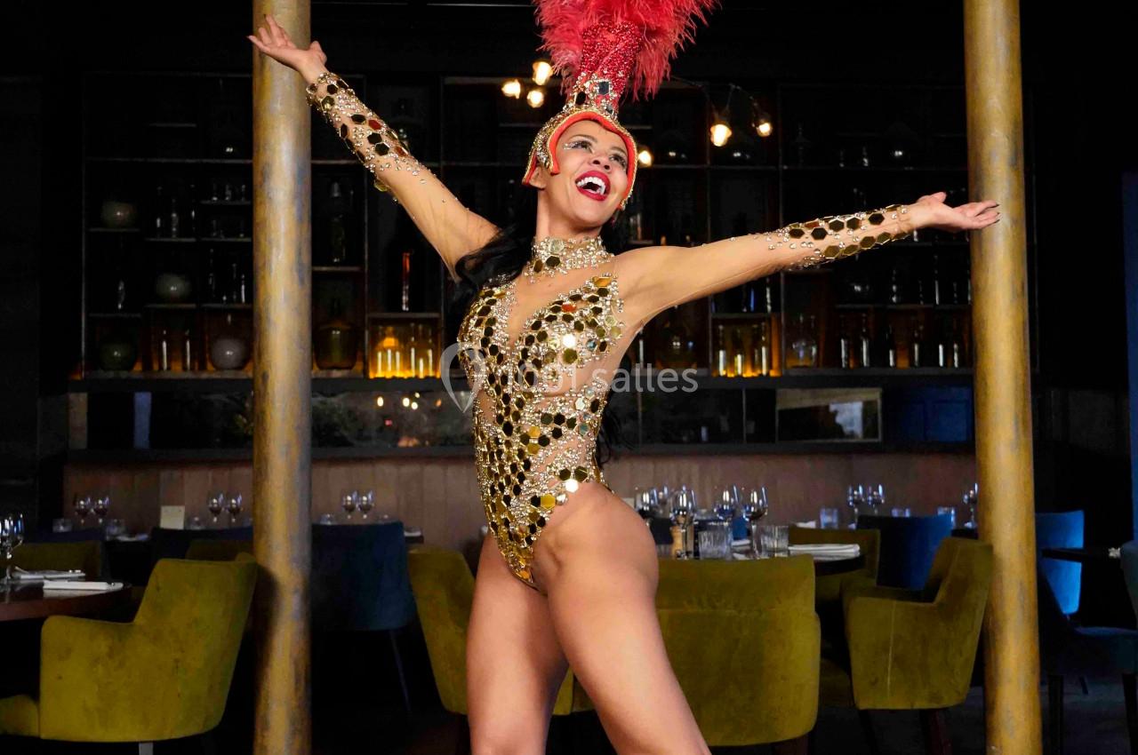 Danseuse en costume scintillant avec coiffe à plumes rouges, posant dans un restaurant élégant avec des tables dressées.