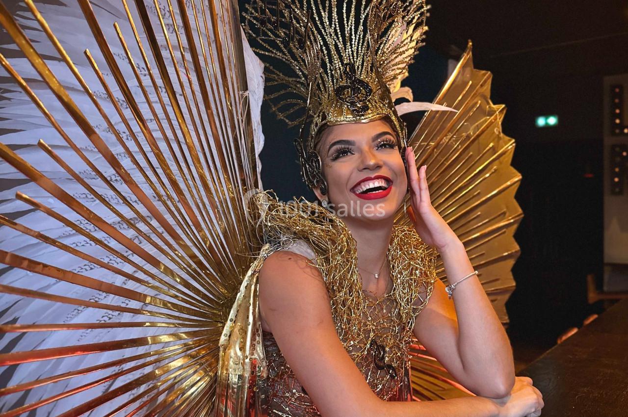 Personne souriante portant un costume doré avec des ailes et une coiffe élaborée, appuyée sur un comptoir.