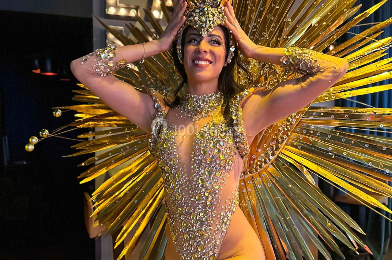 Danseuse en tenue scintillante dorée avec un costume à plumes et un diadème, posant dans un décor intérieur.