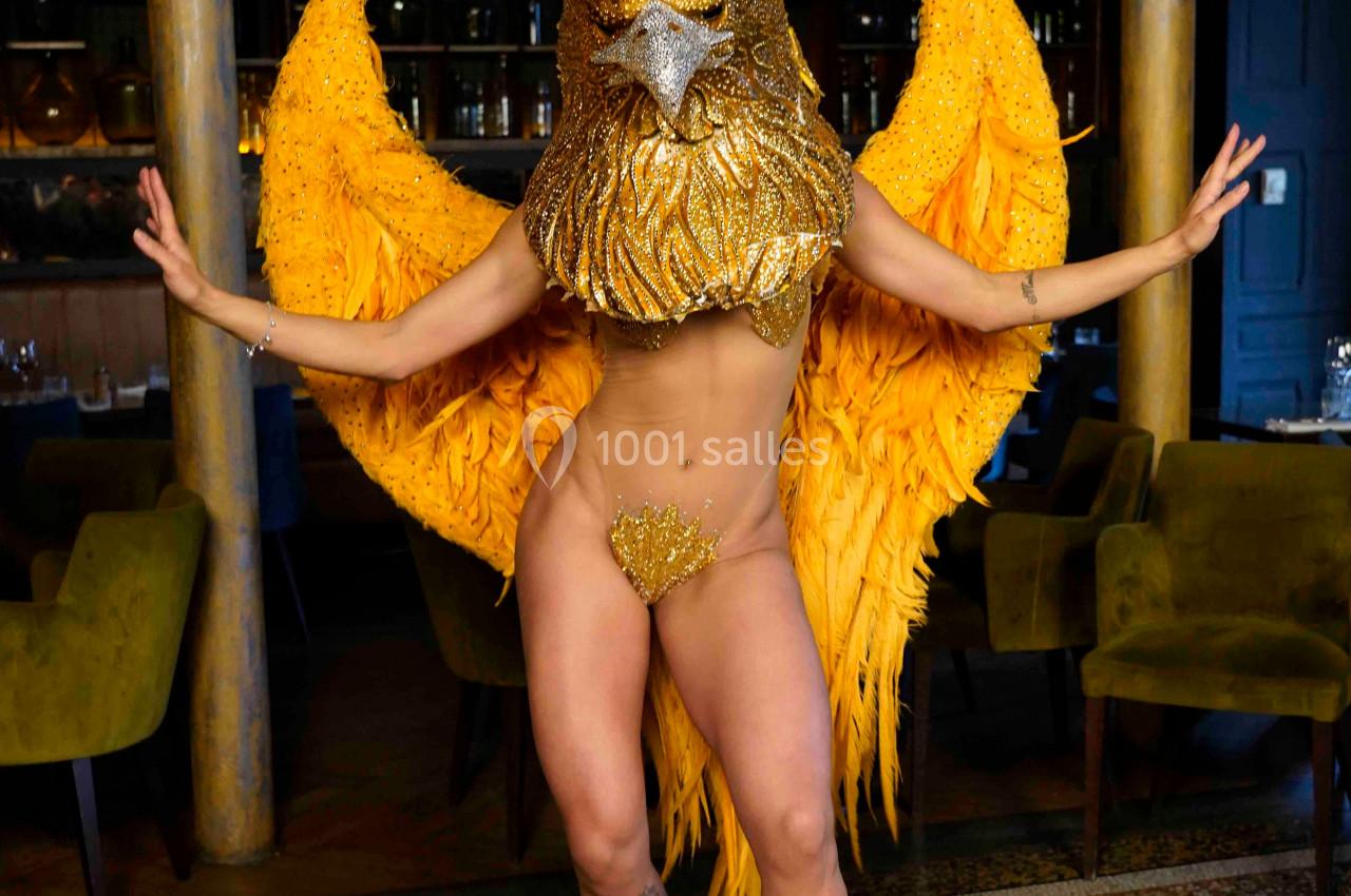 Personne portant un costume doré avec des ailes et un masque en forme de tête d'oiseau, posant dans un intérieur élégant.