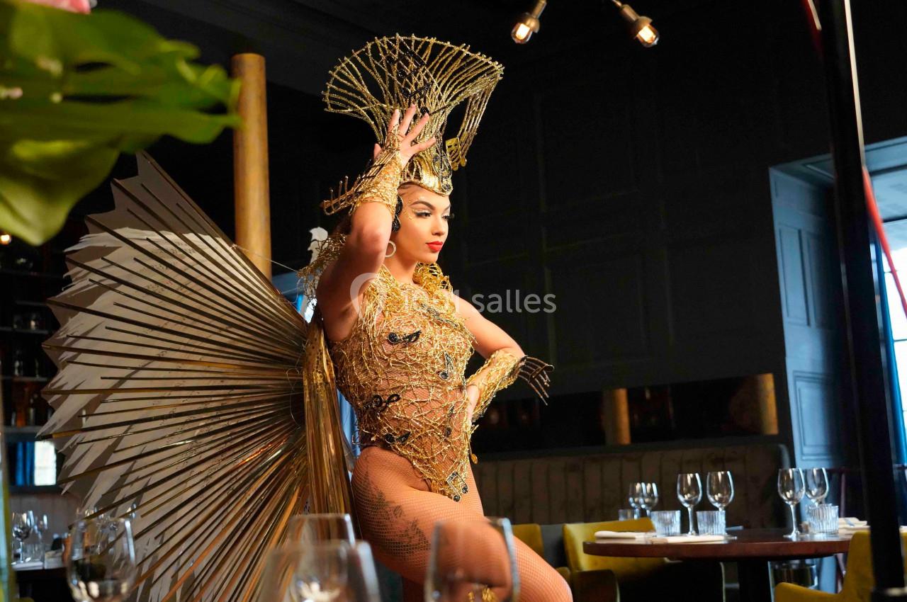 Danseuse en costume doré élaboré avec plumes et ornements, posant dans un restaurant élégant.