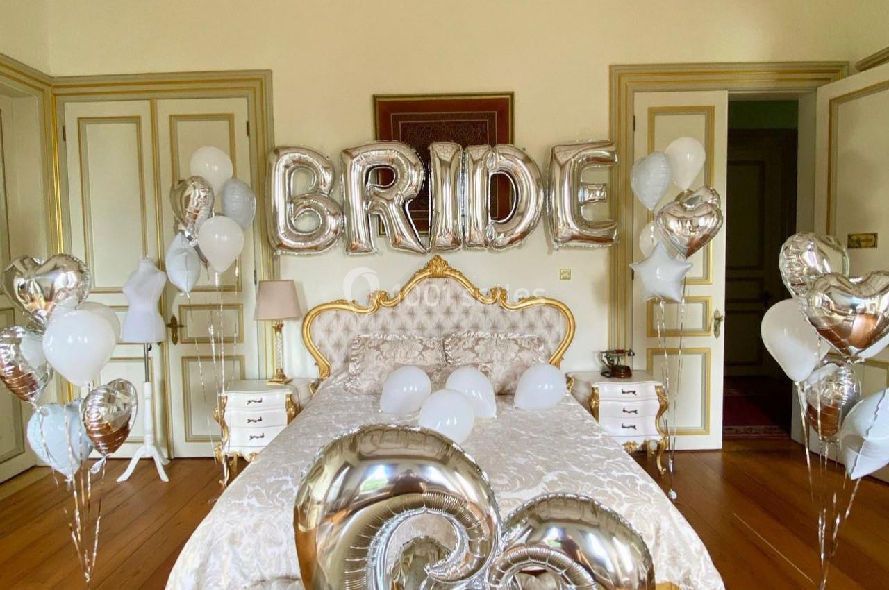 Chambre décorée avec des ballons argentés et blancs, dont des lettres formant ’BRIDE’, au-dessus d'un lit élégant.