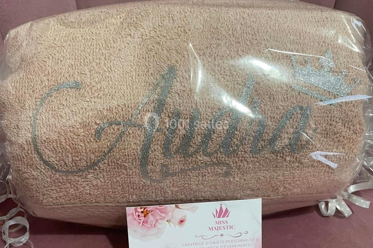 Serviette beige personnalisée avec un prénom brodé et une couronne, emballée dans un plastique transparent sur un fond rose.