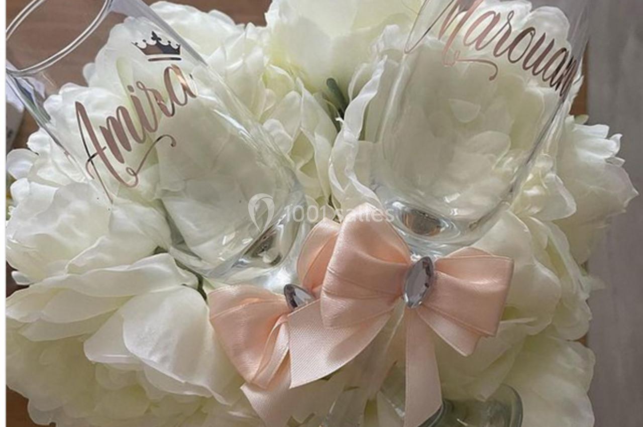 Deux flûtes gravées avec des prénoms reposent sur un bouquet de fleurs blanches, ornées d'un nœud rose pâle.