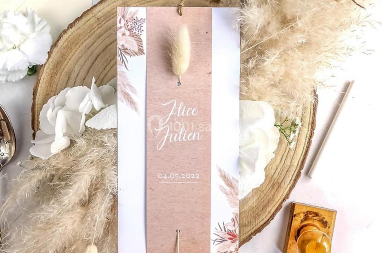 Faire-part de mariage posé sur une tranche de bois, entouré de fleurs blanches, pampas et cachet de cire.