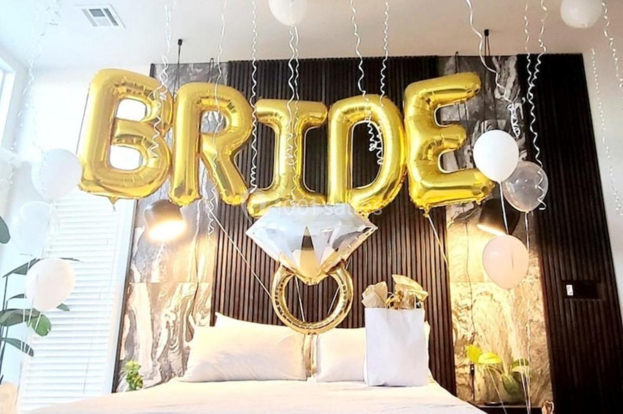 Ballons dorés formant le mot ’BRIDE’ et décorations de mariage au-dessus d'un lit dans une chambre lumineuse.