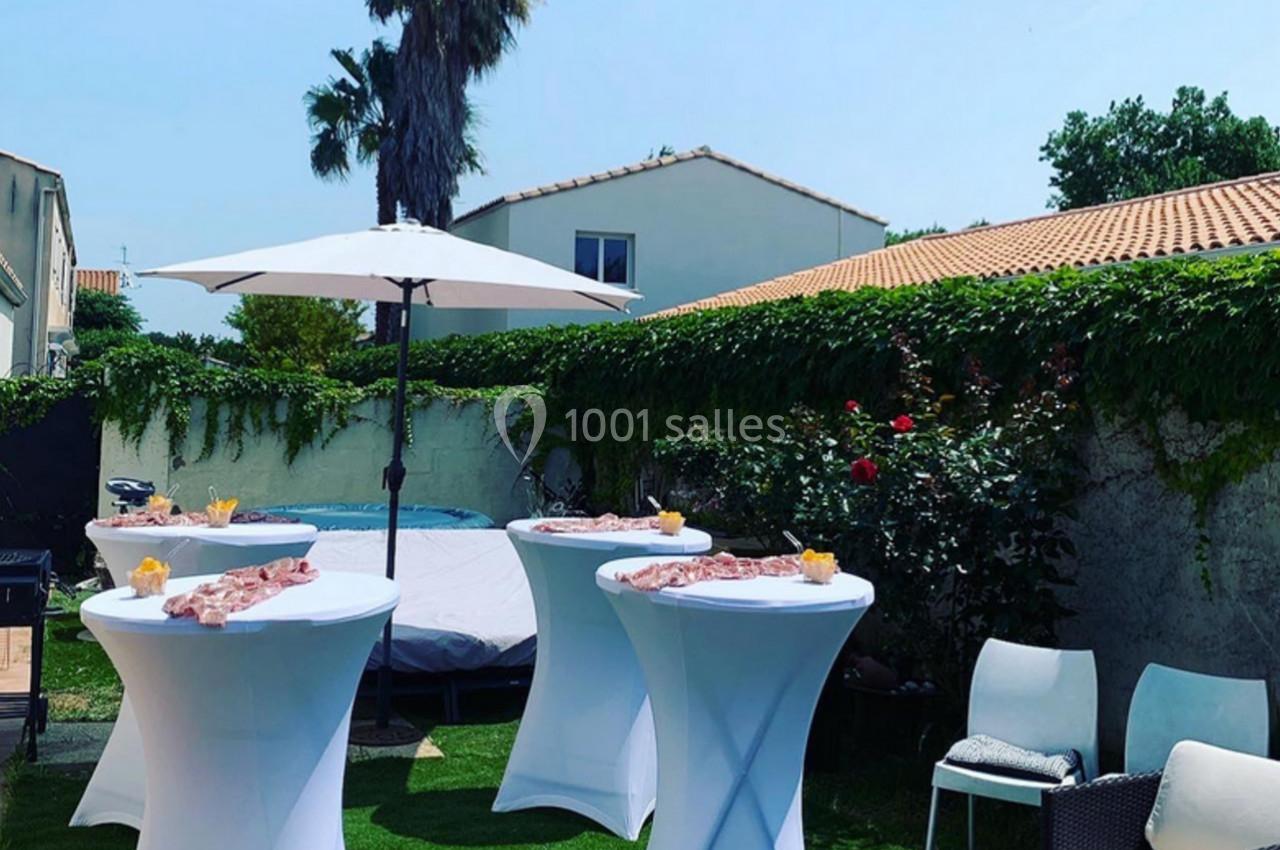 Tables hautes dressées avec des amuse-bouches dans un jardin ensoleillé, entourées de chaises et d'un parasol.