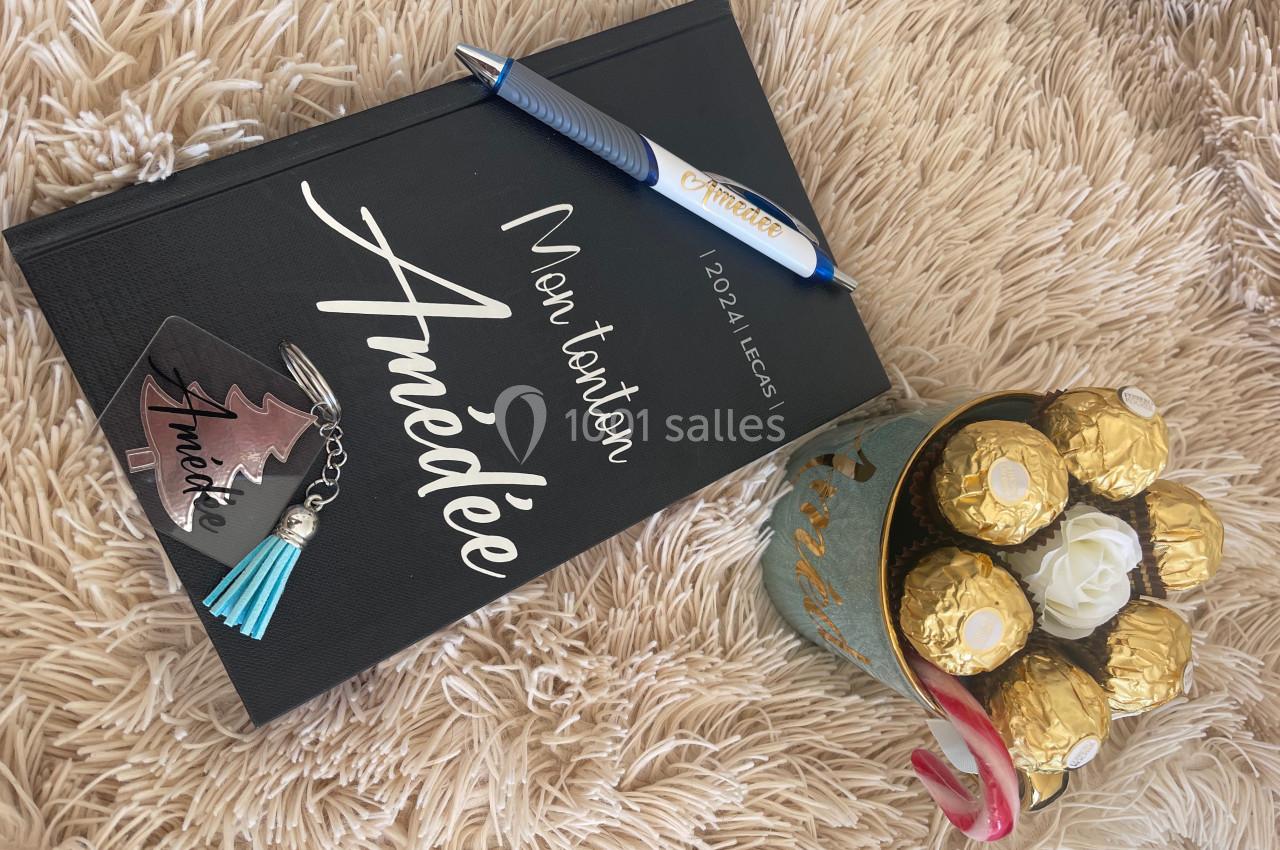 Carnet noir personnalisé, stylo, porte-clés et bouquet de chocolats sur un plaid beige.