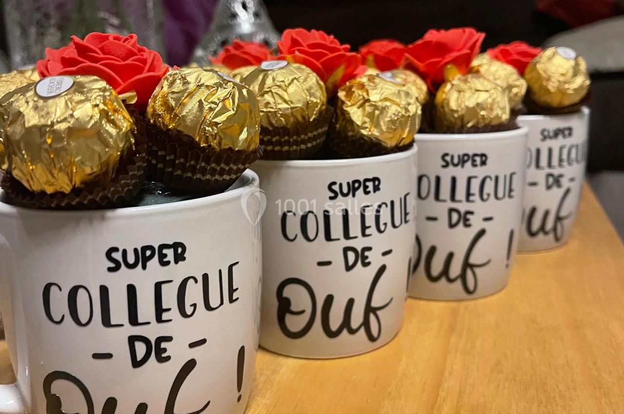 Tasses blanches avec l'inscription ’Super collègue de ouf !’ remplies de chocolats dorés et décorées de fleurs rouges.
