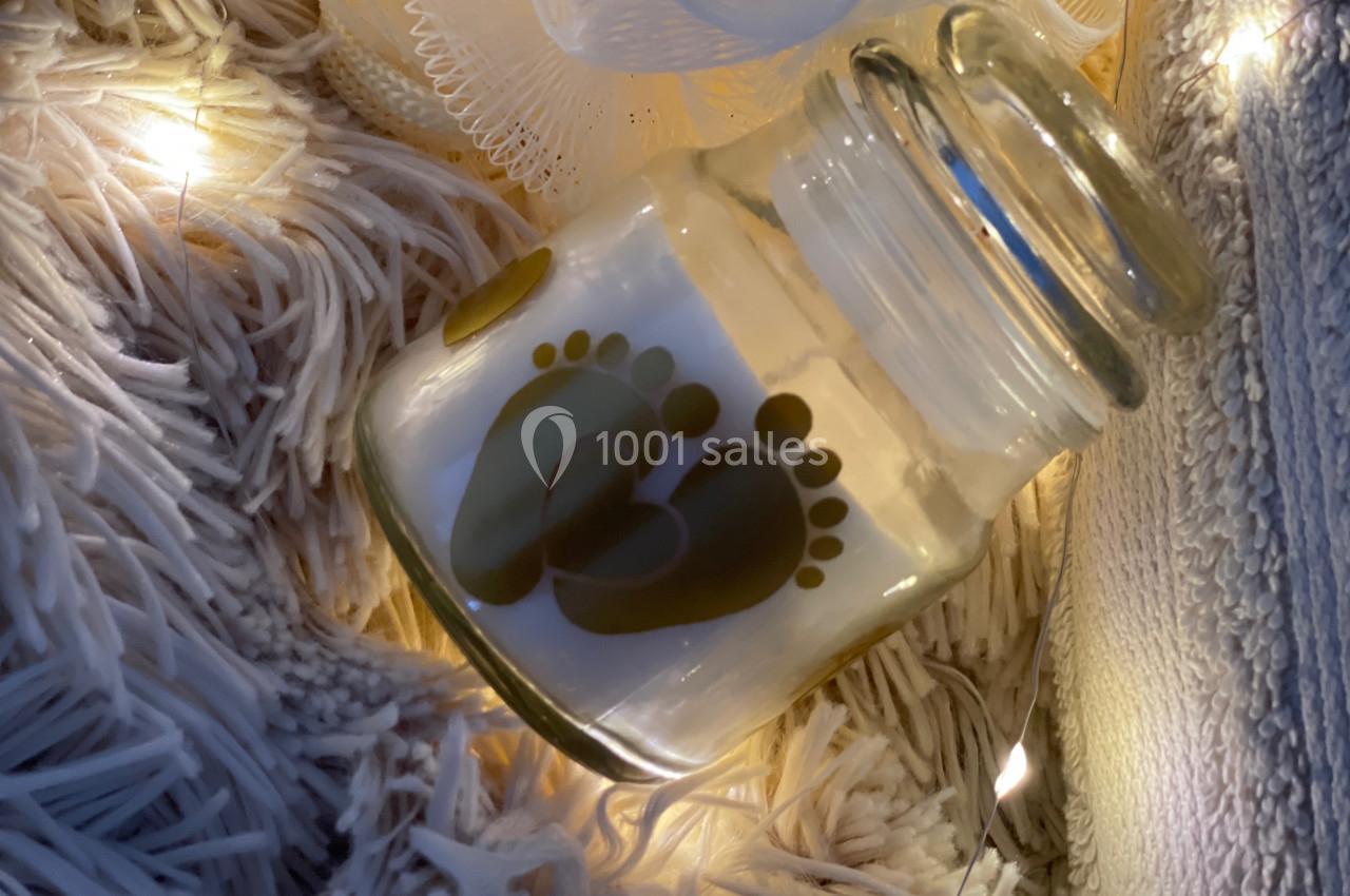 Bougie dans un pot en verre décoré d'empreintes de pieds, posée sur un tapis texturé avec des lumières douces.