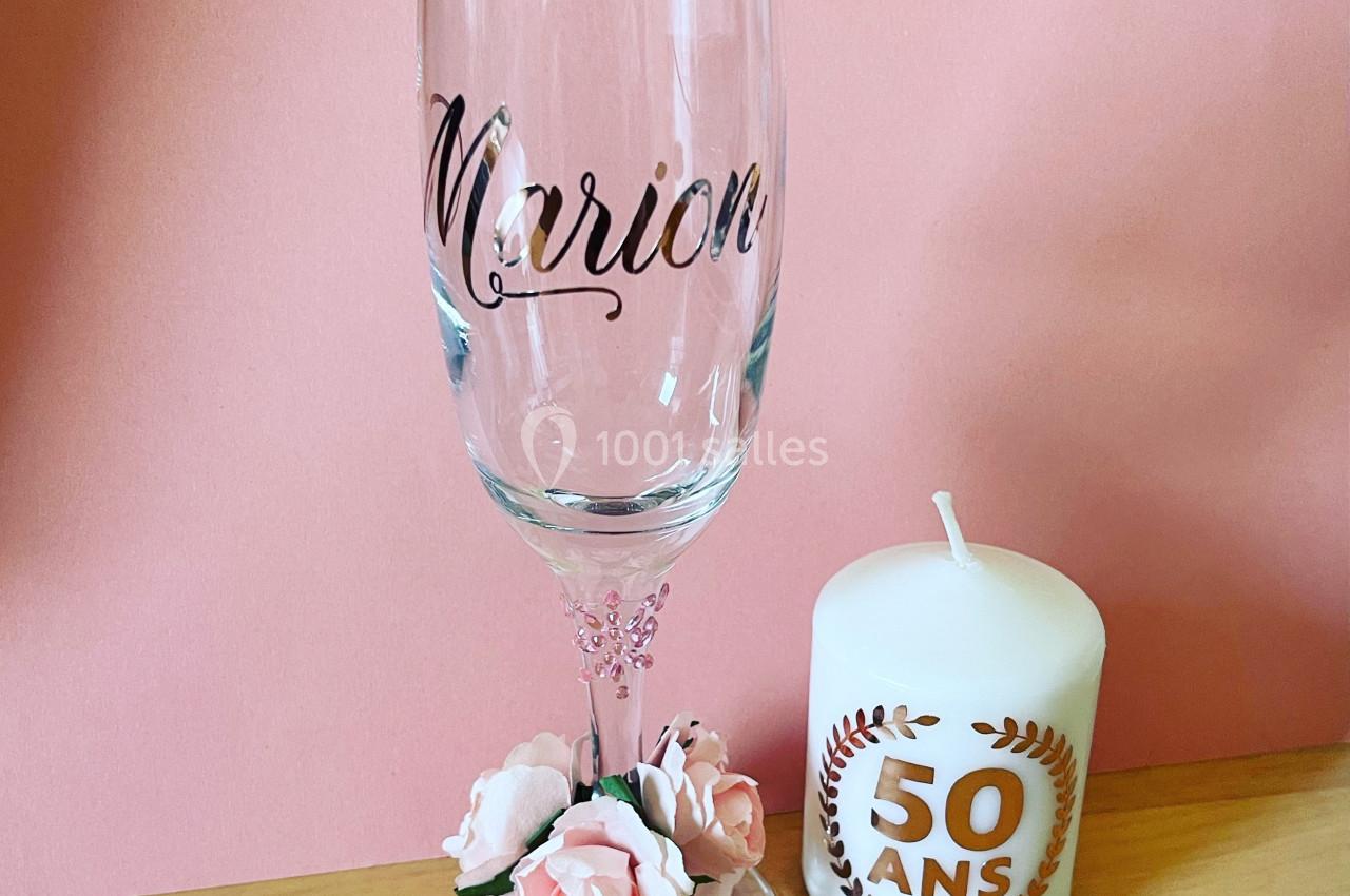 Flûte personnalisée avec le prénom ’Marion’ et bougie marquée ’50 ans’, posées sur une table devant un fond rose.