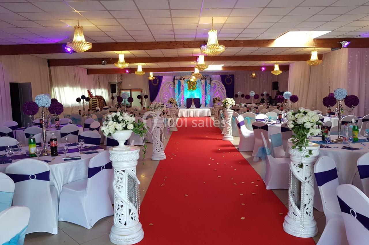 Salle de réception décorée avec des chaises blanches, des tables dressées, un tapis rouge et des ornements floraux.