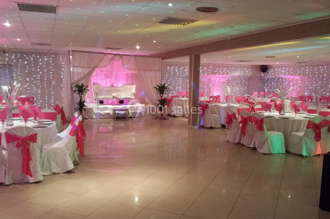 Salle de réception décorée avec des nappes blanches, des nœuds roses sur les chaises et un éclairage tamisé.