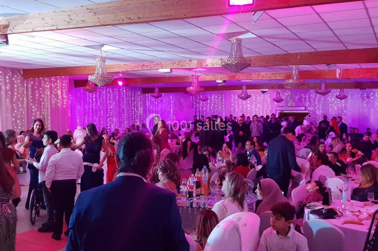 Salle de réception animée avec de nombreuses personnes assises et debout, éclairée par des lumières roses.