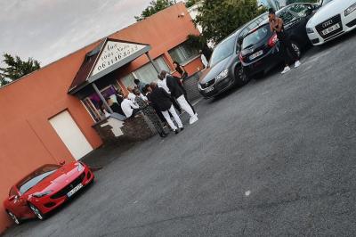 Plusieurs personnes discutent devant un bâtiment avec des voitures garées, dont une voiture rouge sportive.