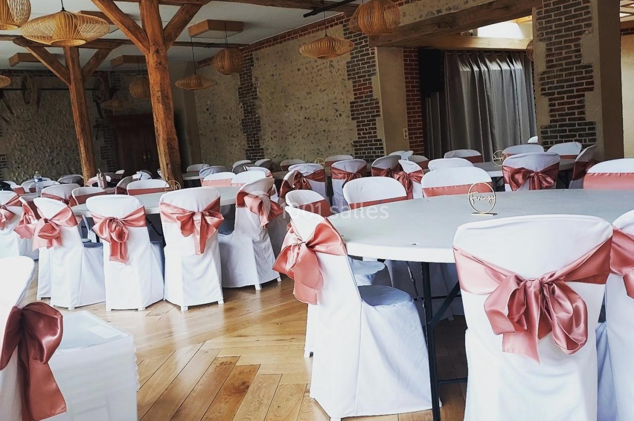 Salle de réception avec tables rondes et chaises blanches décorées de nœuds roses, parquet en bois clair et poutres…