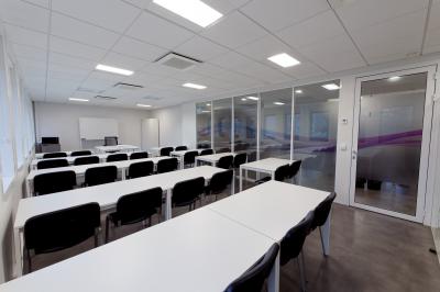 Salle de formation lumineuse avec tables blanches, chaises noires et grandes fenêtres laissant entrer la lumière naturelle.