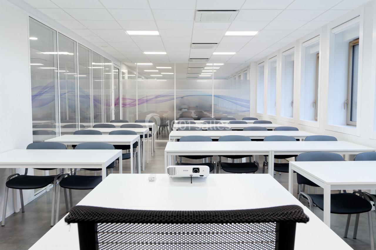 Salle de classe moderne et lumineuse avec des rangées de tables et chaises, vue depuis un bureau équipé d’un projecteur.