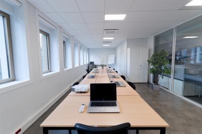 Salle de formation lumineuse avec tables blanches, chaises noires et grandes fenêtres laissant entrer la lumière naturelle.
