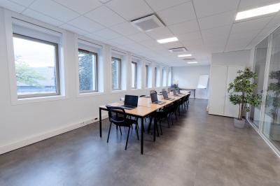 Salle de formation lumineuse avec tables blanches, chaises noires et grandes fenêtres laissant entrer la lumière naturelle.
