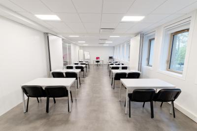 Salle de formation lumineuse avec tables blanches, chaises noires et grandes fenêtres laissant entrer la lumière naturelle.