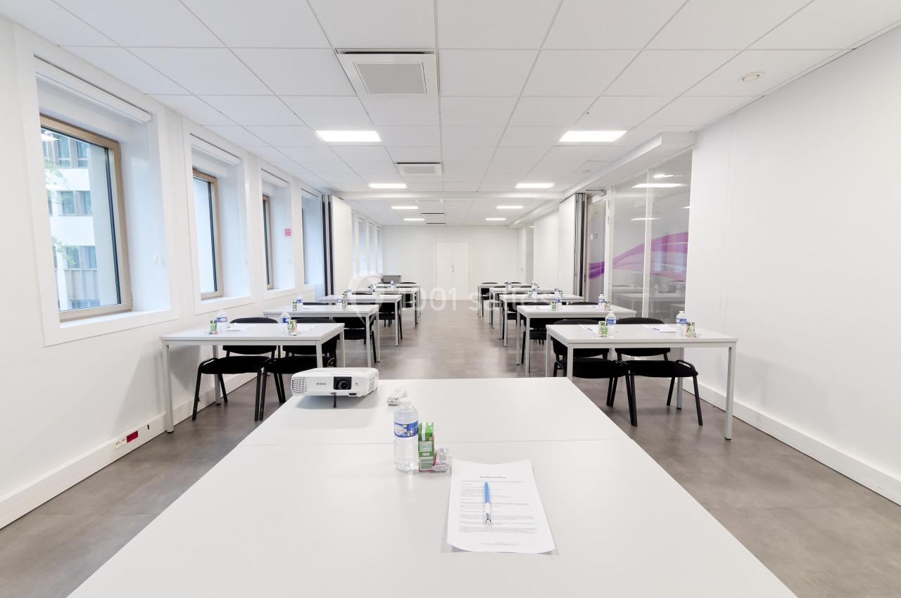 Salle de formation lumineuse avec des tables alignées, des chaises, des documents et une bouteille d'eau sur chaque table.