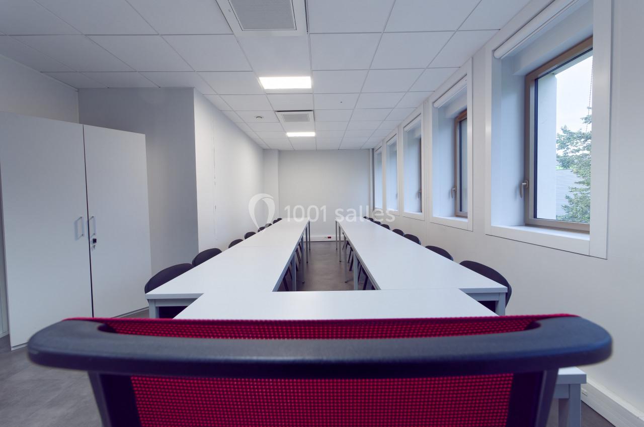 Salle de réunion vide avec tables blanches disposées en U, chaises noires et fenêtres laissant entrer la lumière naturelle.