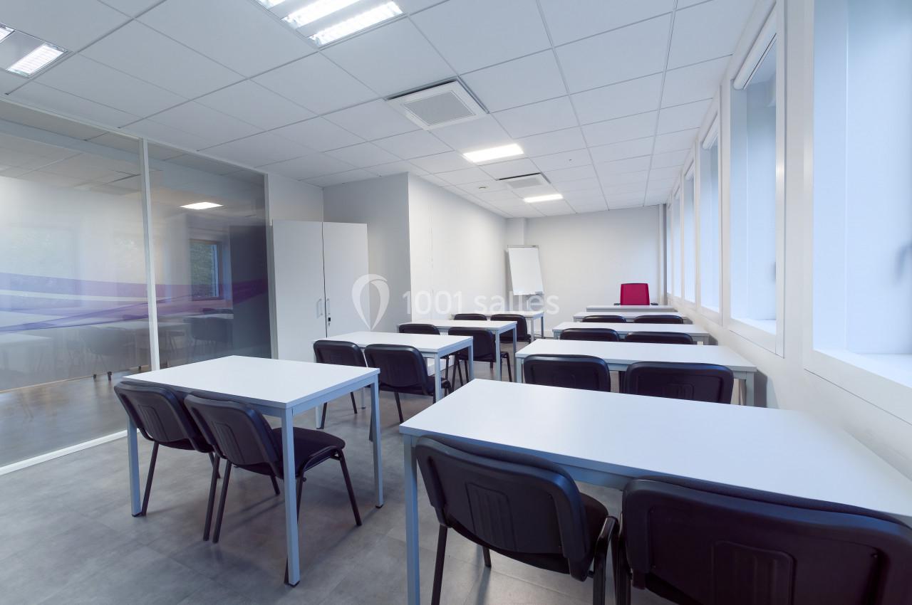 Salle de classe vide avec des tables, des chaises noires, un tableau blanc et de grandes fenêtres laissant entrer la lumière.