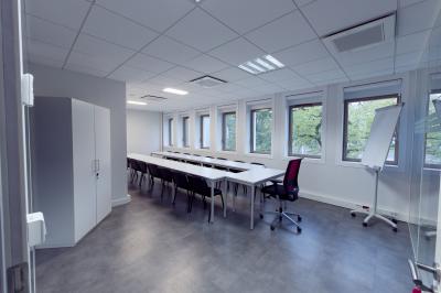 Salle de formation lumineuse avec tables blanches, chaises noires et grandes fenêtres laissant entrer la lumière naturelle.