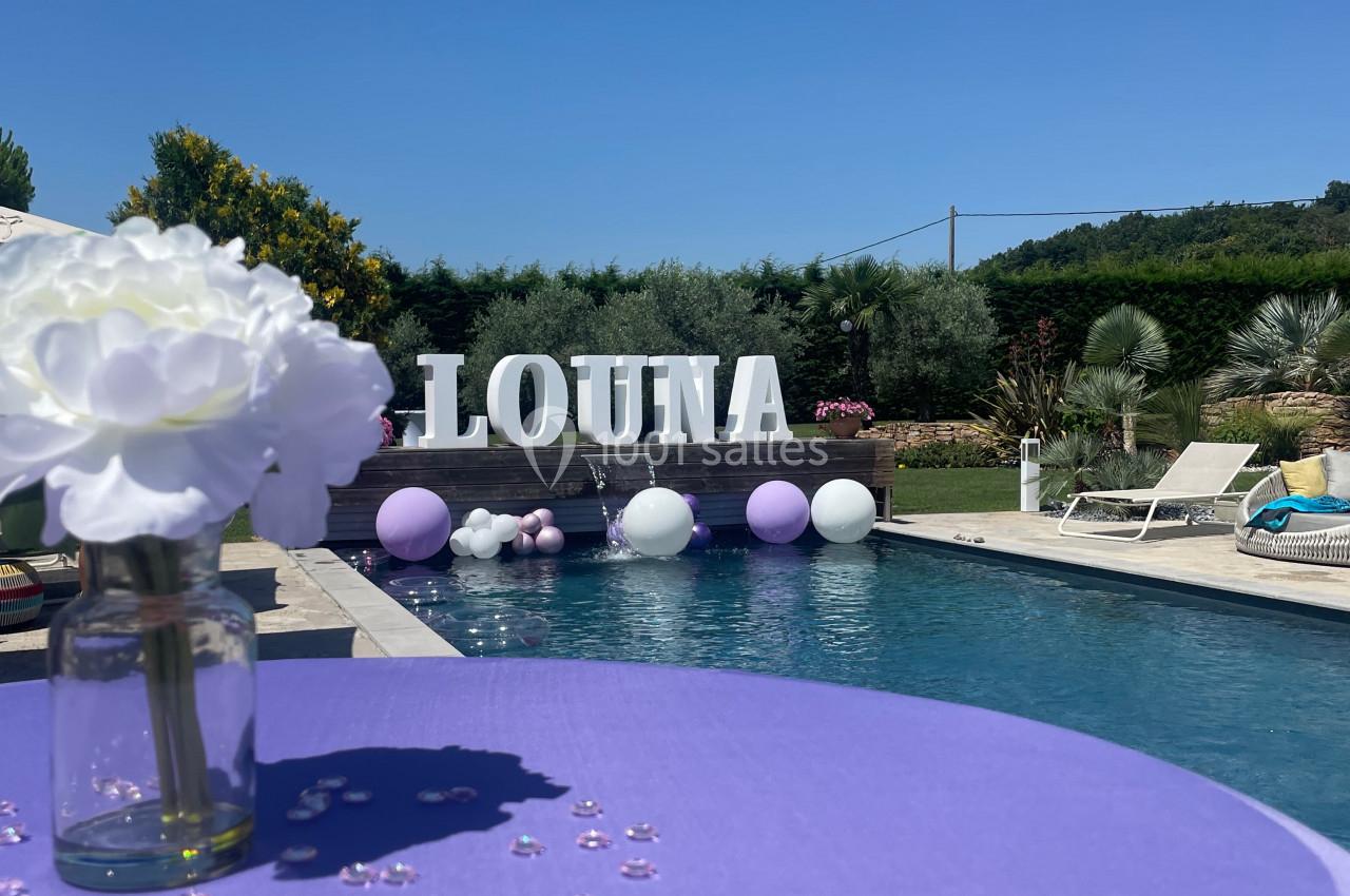 Table violette décorée de perles et fleurs blanches, près d'une piscine entourée de ballons et d'une végétation estivale.