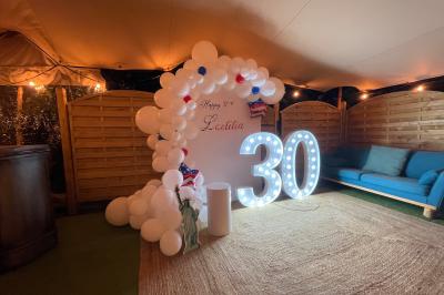 Décoration d'anniversaire avec ballons violets et blancs, chiffre lumineux ’18’ et présentoirs cylindriques blancs.