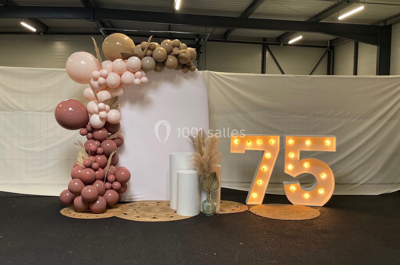 Décoration avec arche de ballons dans des tons neutres et chiffre lumineux ’75’ dans un espace intérieur.