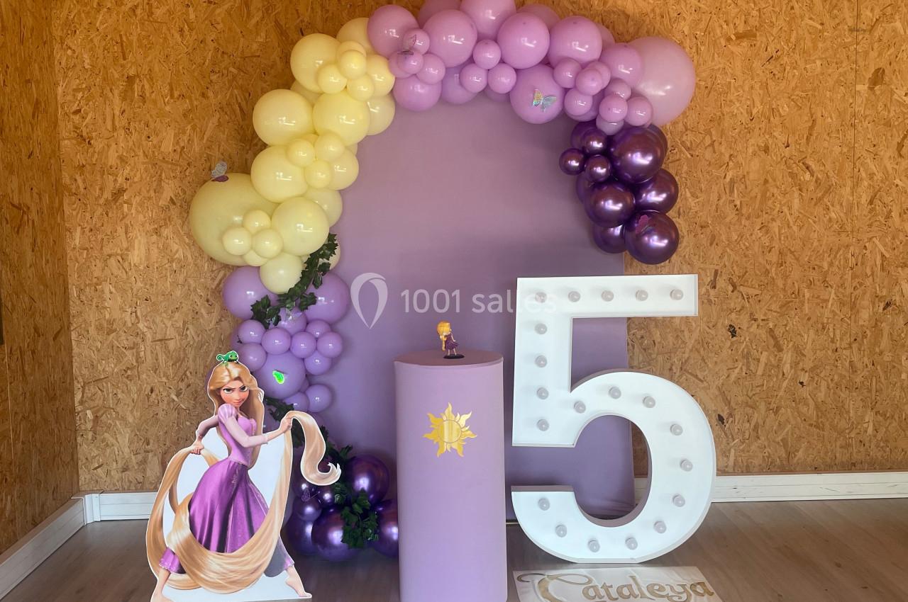 Décoration d'anniversaire avec ballons violets et jaunes, chiffre 5 lumineux, et figurine de princesse devant un mur en bois.