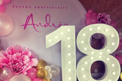 Décoration d'anniversaire avec ballons violets et blancs, chiffre lumineux ’18’ et présentoirs cylindriques blancs.