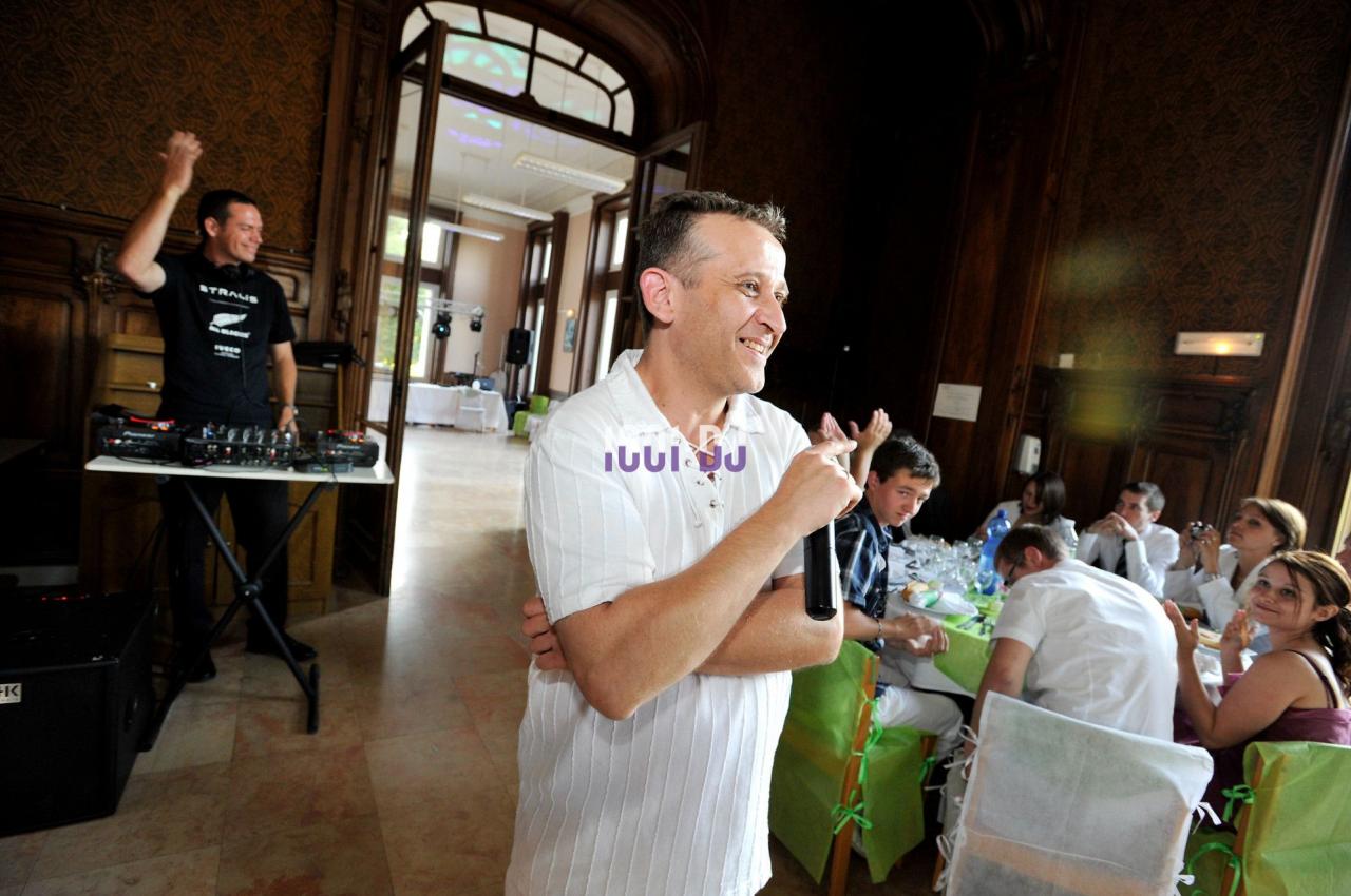 Un homme tenant un micro sourit devant un groupe de personnes assises, avec un DJ en arrière-plan.