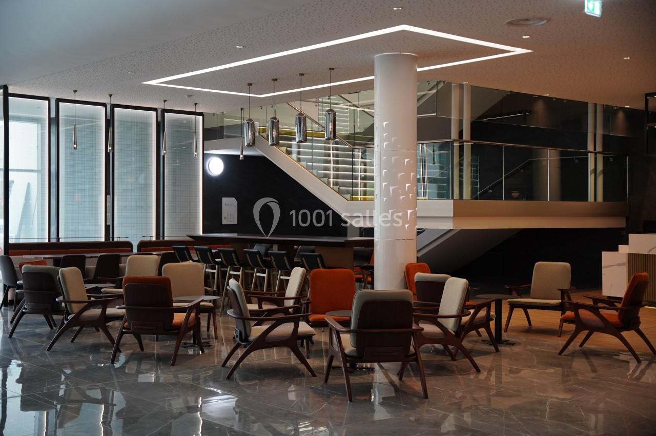 Espace intérieur moderne avec des fauteuils disposés autour de tables, un escalier et un bar en arrière-plan.