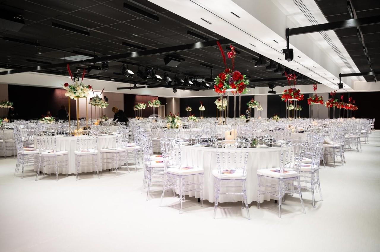 Salle de réception élégante avec tables rondes décorées de fleurs blanches et rouges sous un éclairage moderne.