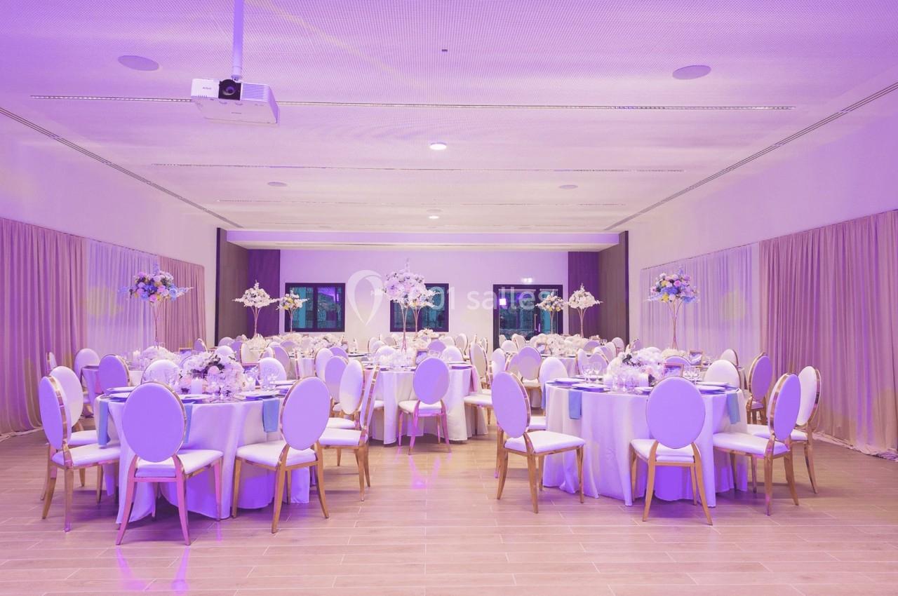 Salle de réception décorée avec des tables rondes, nappes blanches, chaises élégantes et éclairage violet.