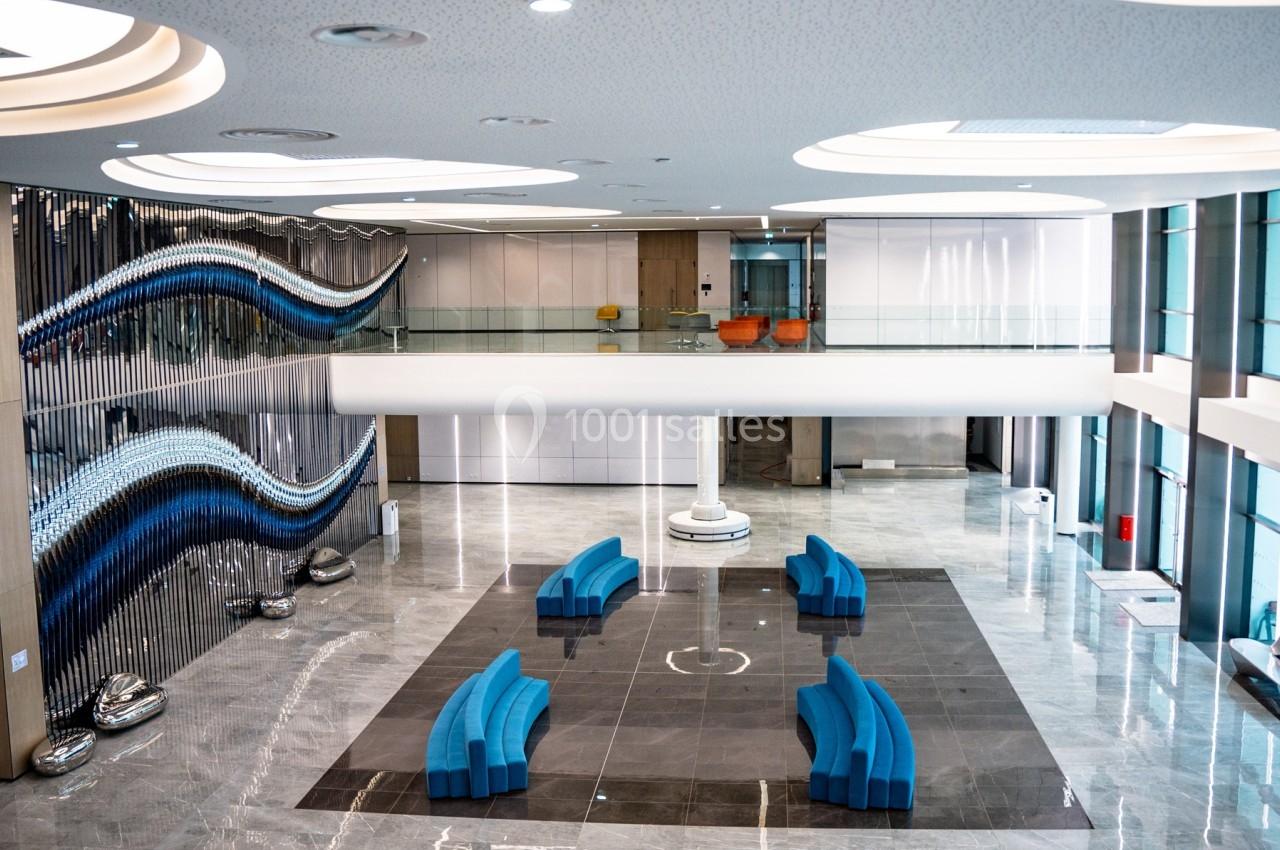 Hall moderne avec sol en marbre, sièges bleus courbés, sculptures murales ondulées et mezzanine vitrée.