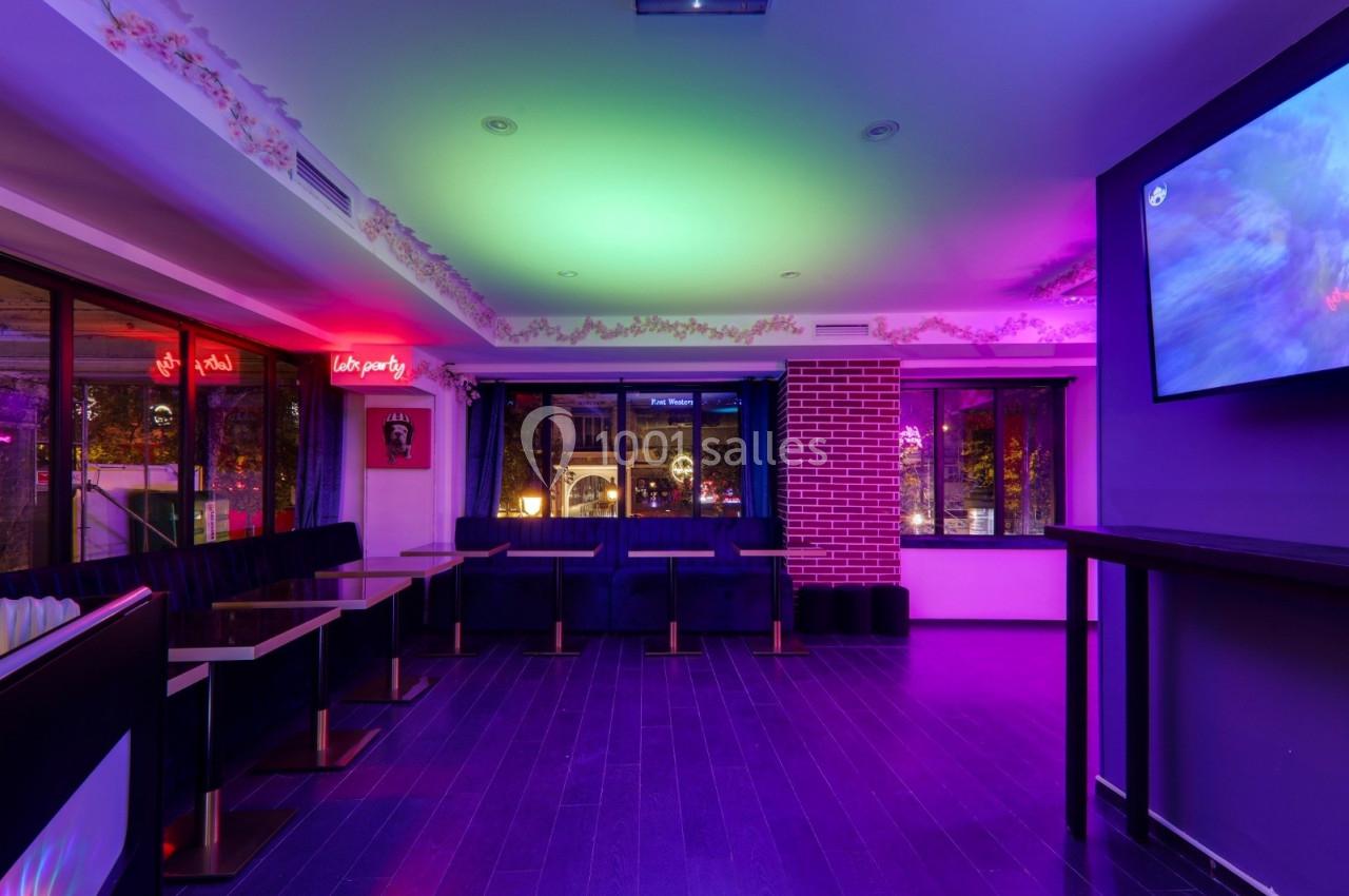 Salle lumineuse avec éclairage coloré, banquettes noires, tables en bois et grandes fenêtres donnant sur l'extérieur.