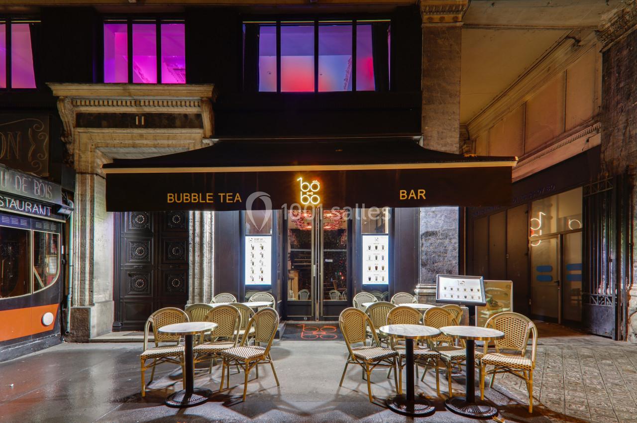 Façade d'un bar à bubble tea avec terrasse extérieure, tables et chaises en rotin sous un auvent noir.