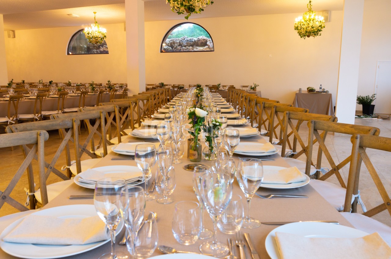 Salle de réception avec longues tables dressées, chaises en bois, vaisselle élégante et décorations florales.