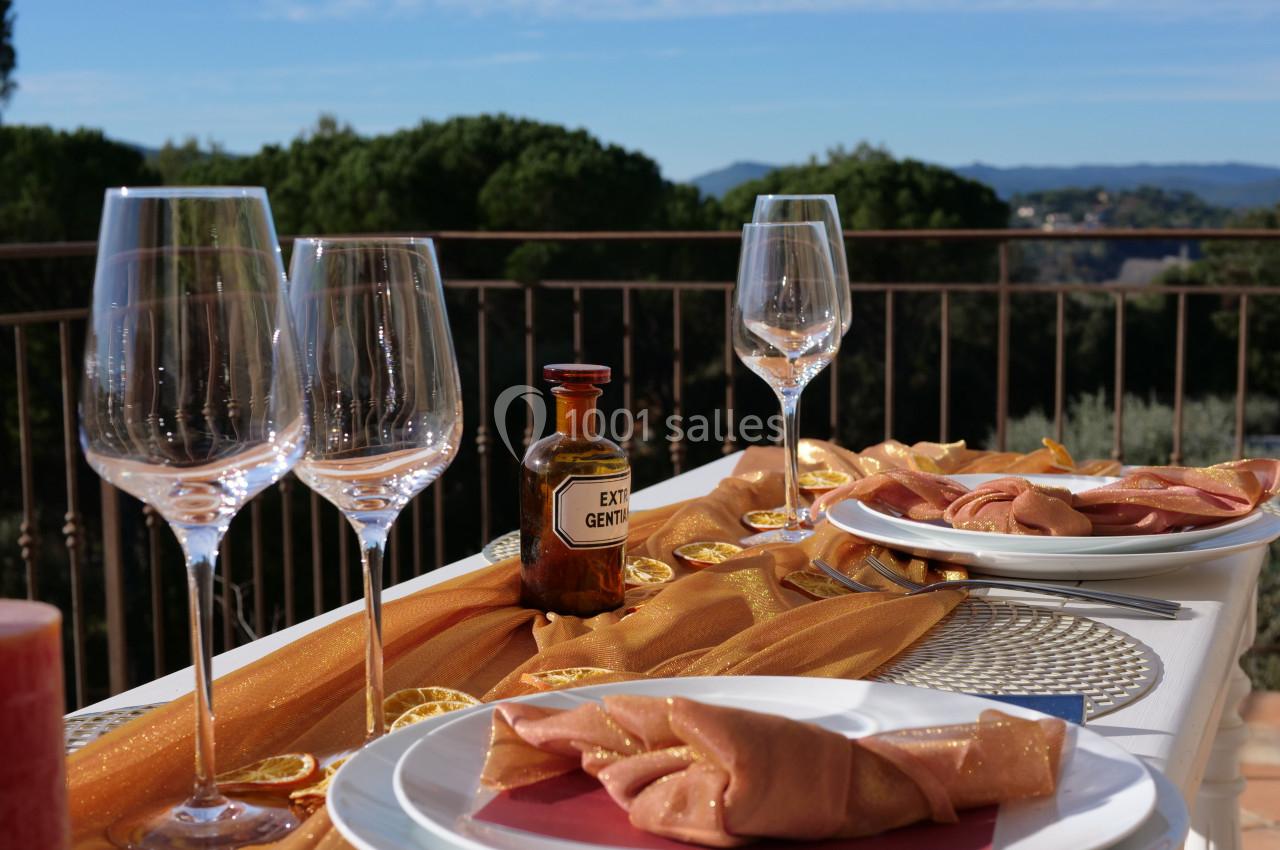 Table dressée en extérieur avec nappage doré, verres à vin, assiettes et bouteille d'huile, vue sur un paysage boisé.