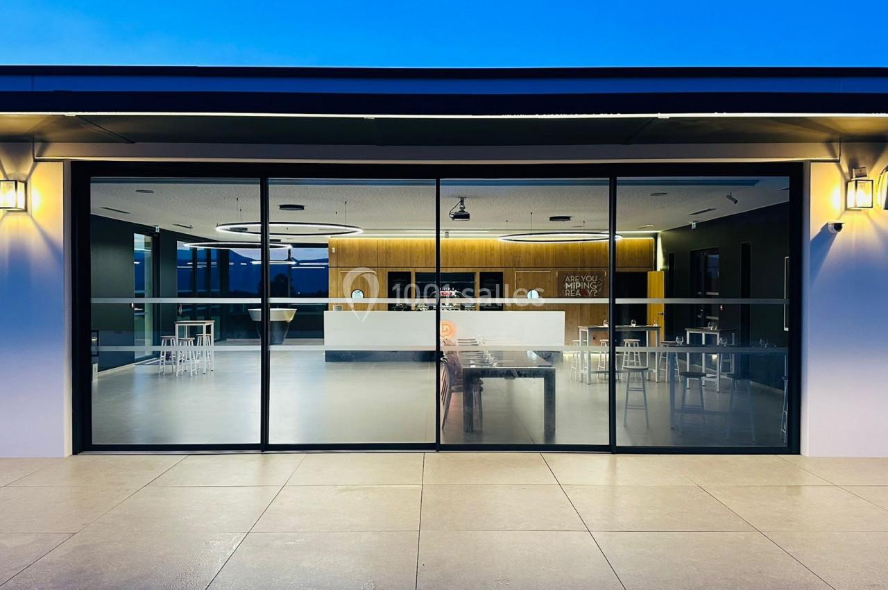 Façade vitrée d'un bâtiment moderne éclairé, donnant sur un espace intérieur avec mobilier minimaliste.