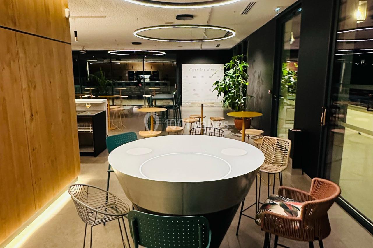 Salle moderne avec table ronde centrale, chaises variées, éclairage circulaire et grandes baies vitrées.